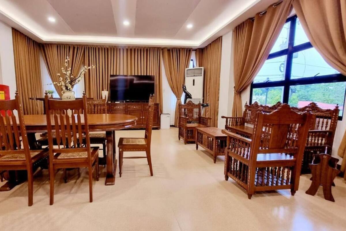Un coin repas avec une table ronde, des chaises en bois, un téléviseur et une grande fenêtre avec des rideaux.