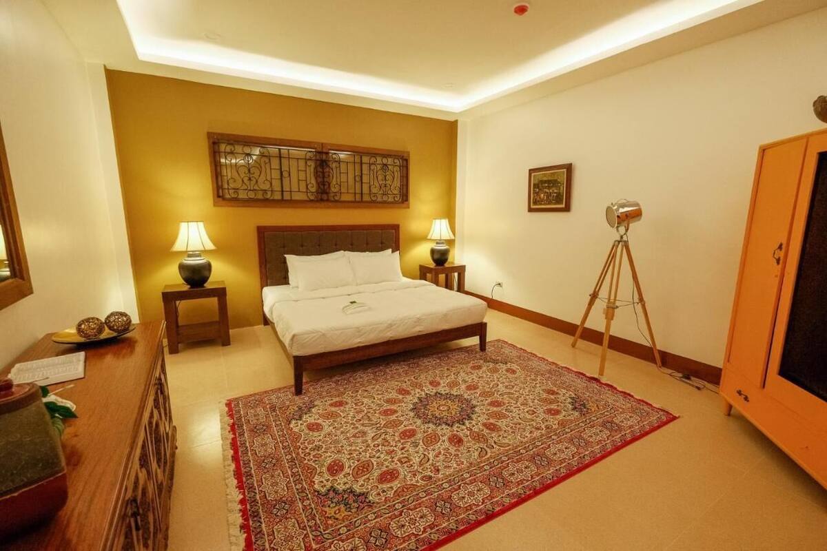 Une chambre d’hôtel comprenant un lit, deux lampes de chevet, une tête de lit en bois, un tapis à motifs, un lampadaire sur trépied, une commode en bois et un meuble de rangement.