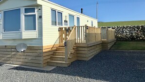 A mobile home with a wooden deck, a satellite dish, and a stone wall in the background.
