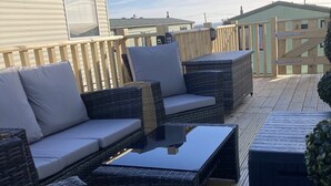 A deck with wicker furniture, including a sofa and ottoman, and a glass-top coffee table.