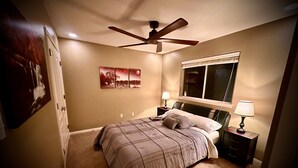 Un dormitorio con cama, ventilador de techo, ventana y mesita de noche con lámpara.