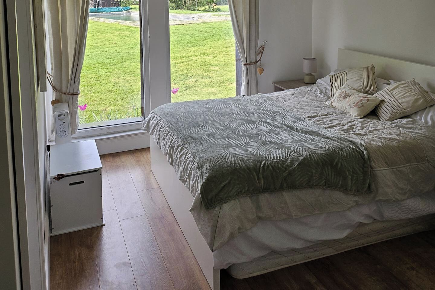 Un dormitorio con cama, cortinas, una mesita de noche y vistas a un jardín verde.