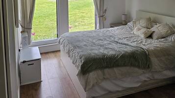 Un dormitorio con cama, cortinas, una mesita de noche y vistas a un jardín verde.