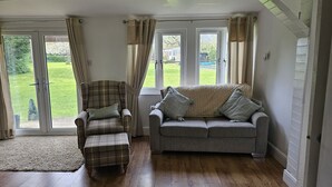 Una habitación con un sillón a cuadros, un sofá gris y piso de madera. Hay dos ventanas con cortinas, una puerta y vistas a un césped verde.