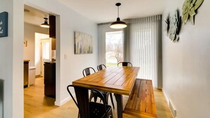 Un coin repas avec une table et des chaises en bois, un luminaire suspendu et une vue sur la cuisine par une porte ouverte.