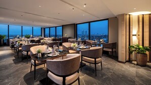A modern dining area with multiple tables and chairs, large windows offering a city view, and a high ceiling with recessed lighting. - CLASSYC Hotel (Hanoi)