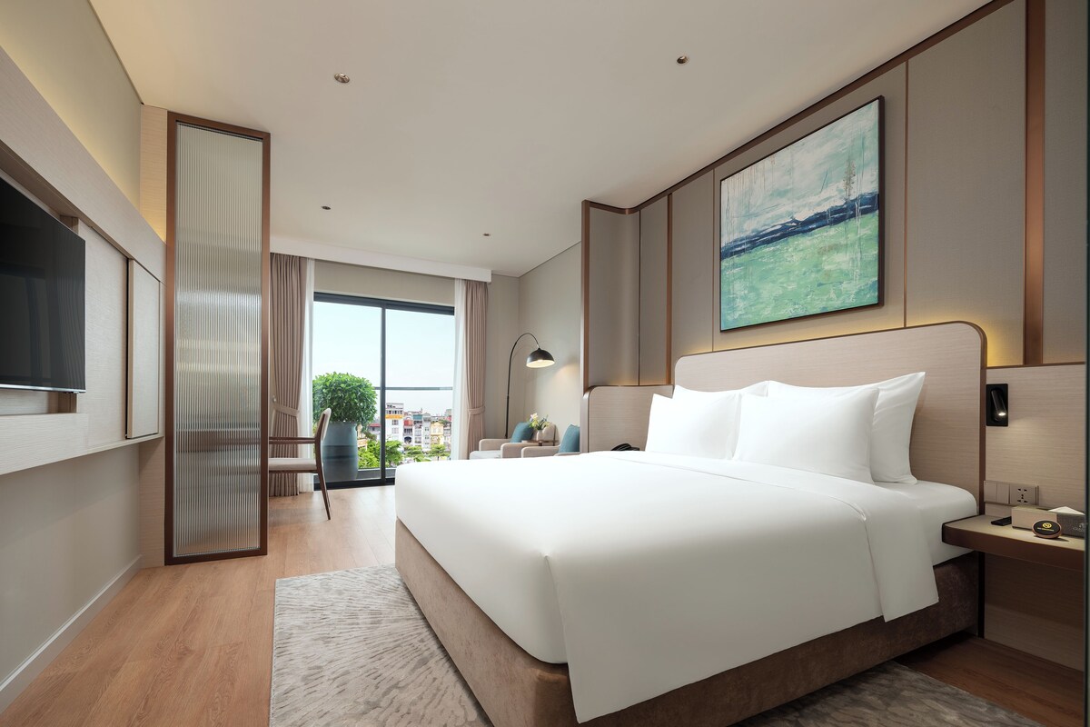 A modern hotel room with a large bed, a TV, a desk, and a view of the city through large windows.