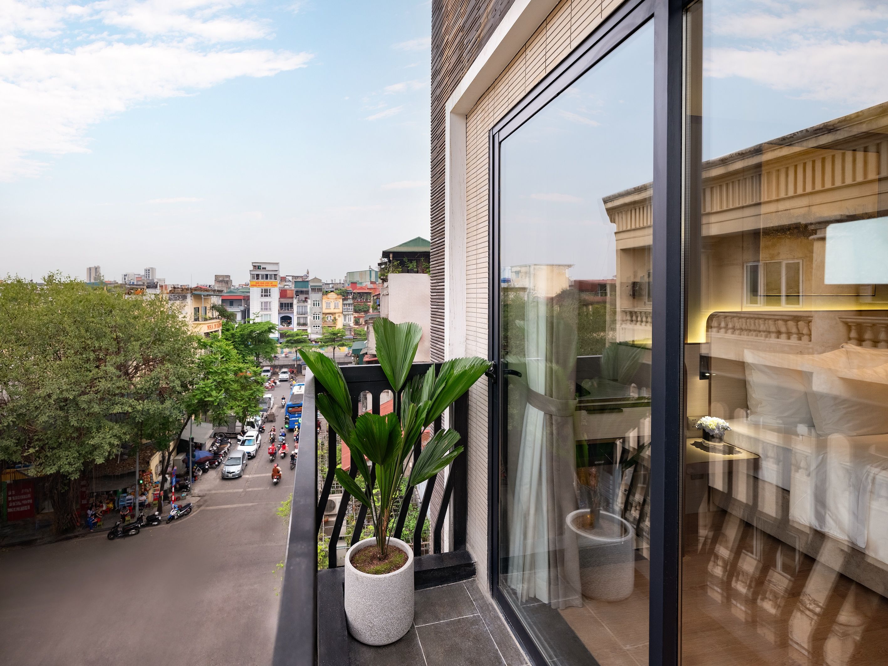 A balcony with a potted plant, a view of a city street, and a reflection of a building.