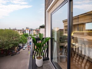 A balcony with a potted plant, a view of a city street, and a reflection of a building. - CLASSYC Hotel (Hanoi)