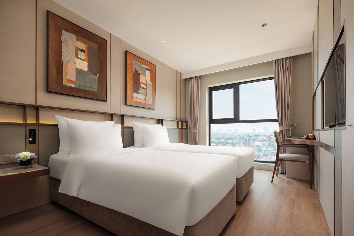 A hotel room with a bed, bedside tables, a desk, and a large window with a city view.