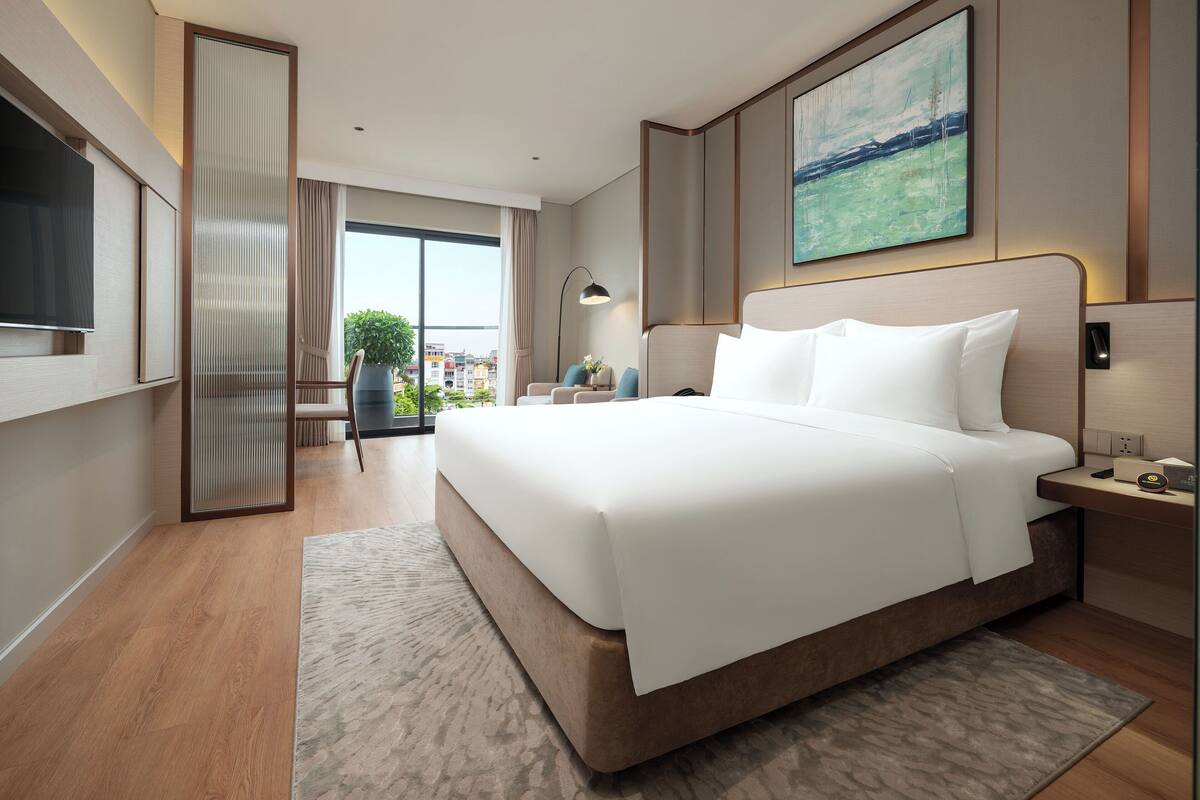 A modern hotel room with a large bed, a TV, a desk, and a view of the city through large windows.