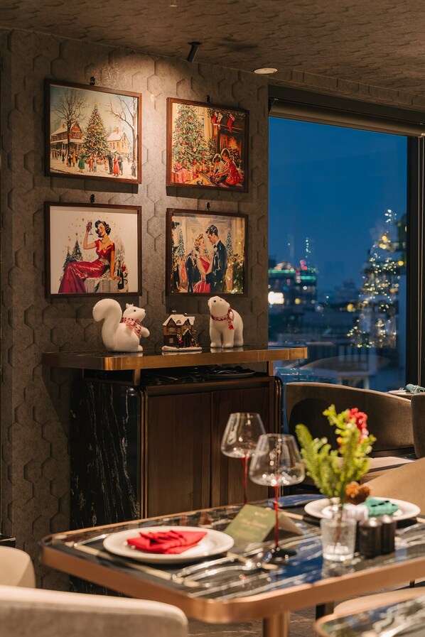 A dining area with a table set for two, a fireplace with decorative items, and a cityscape view through a large window. - CLASSYC Hotel (Hanoi)