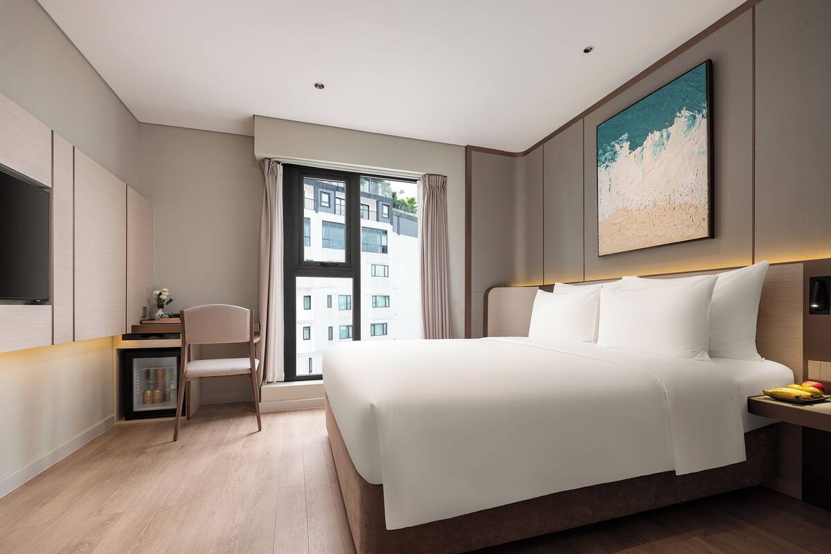 A modern hotel room with a large bed, a desk with a chair, a flat-screen TV, and a window with a view of buildings.