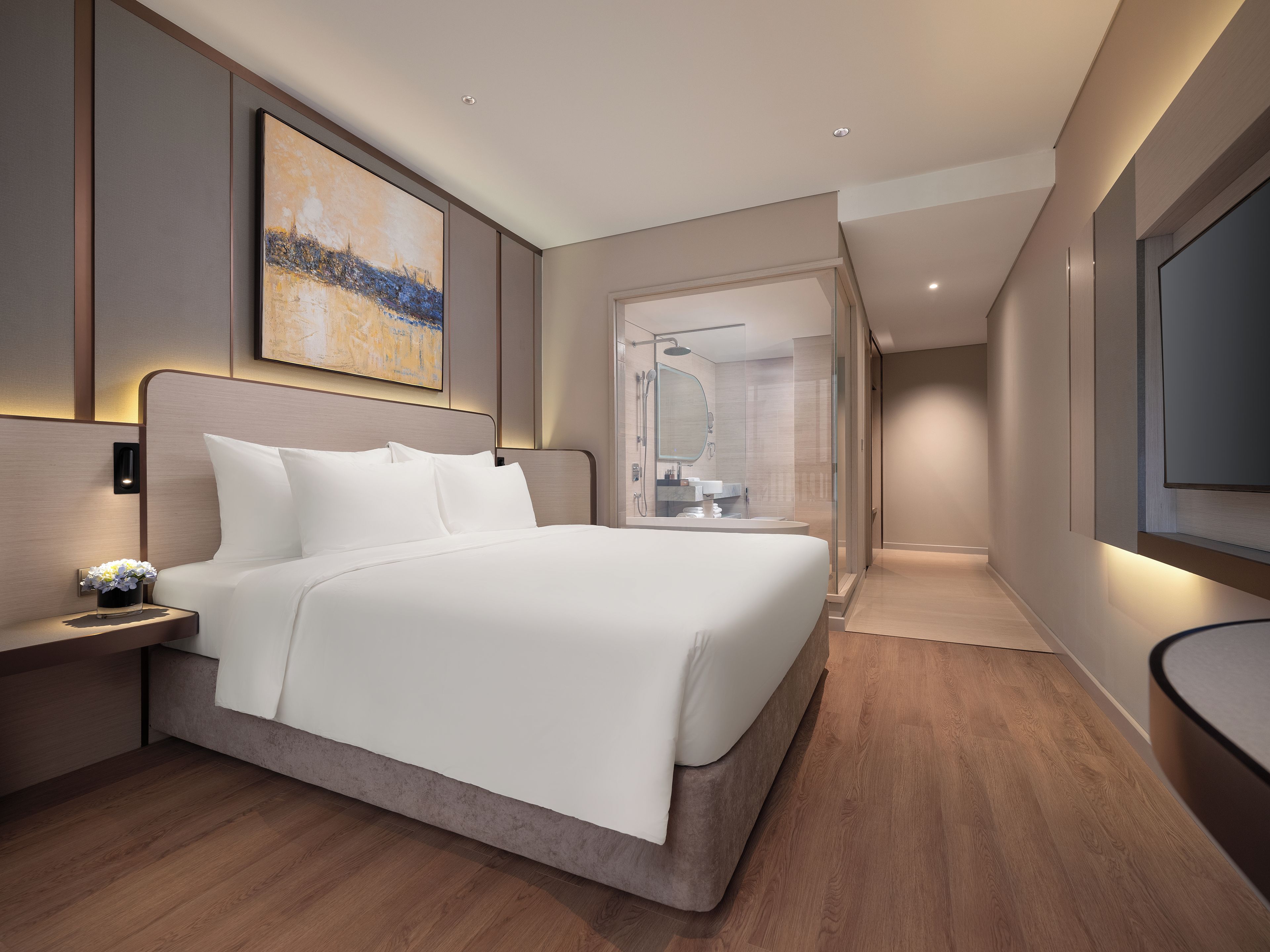 A modern hotel room with a large bed, a bedside table with a vase of flowers, a large painting on the wall, and a TV mounted on the wall.