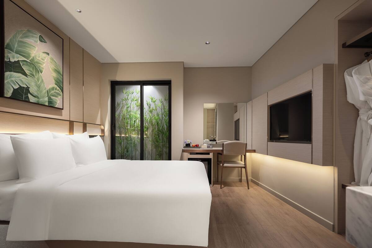 A modern hotel room with a large bed, a desk, a chair, a television, and a large window with a view of greenery.