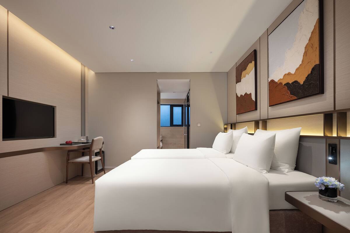 A modern hotel room with a large bed, a desk, a chair, and a flat-screen TV mounted on the wall.