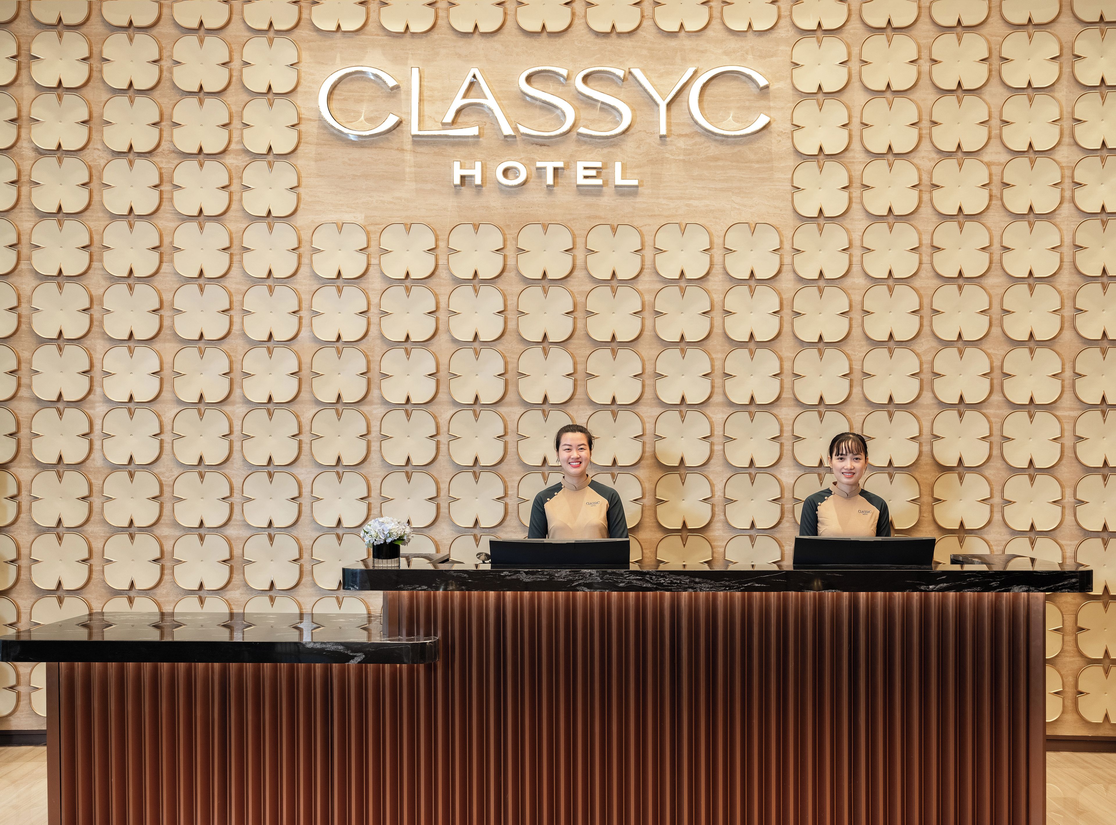 A reception desk at CLASSYHC HOTEL with a patterned wall and two staff members behind it.