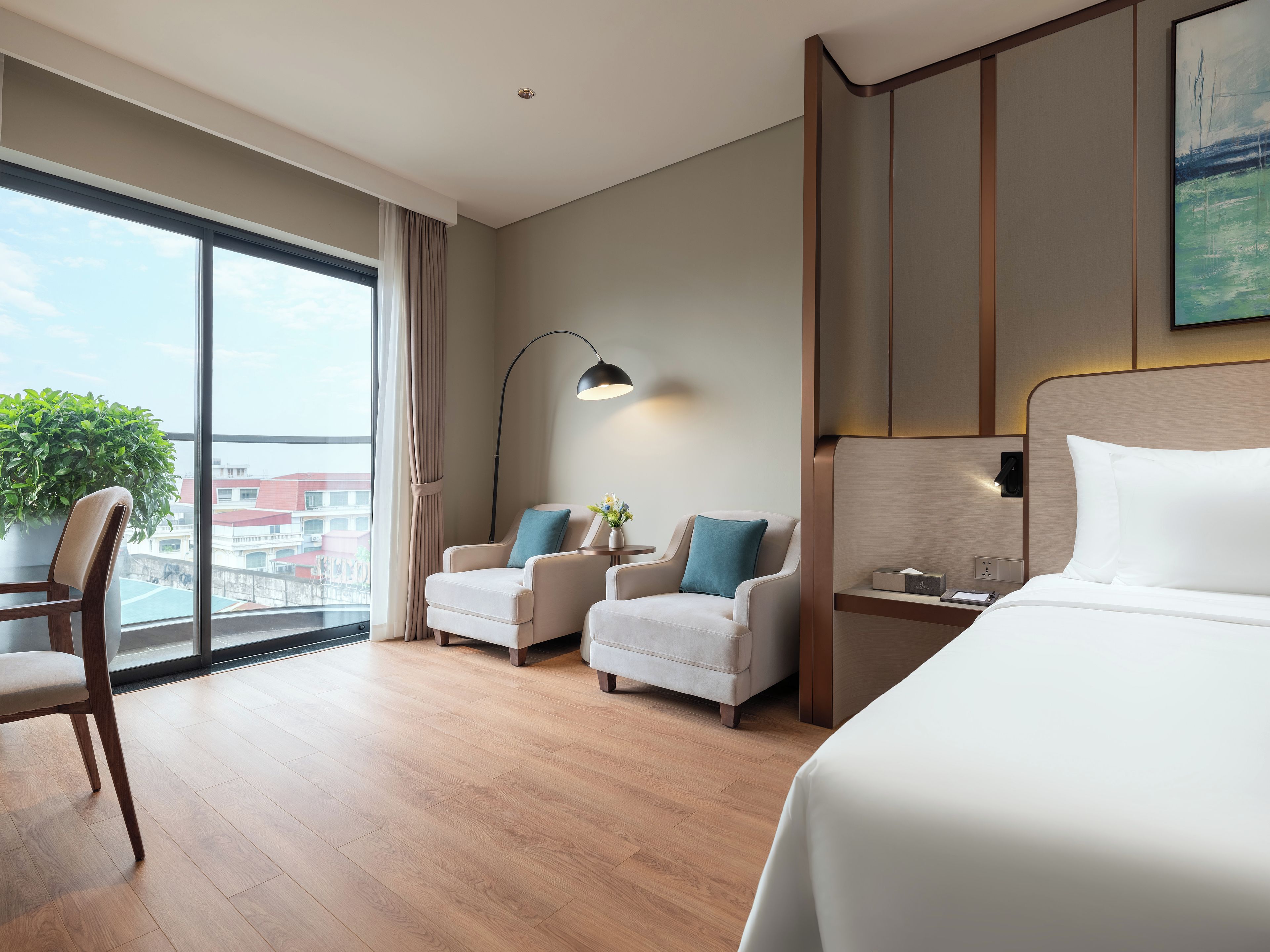 Habitación de hotel con un ventanal grande, dos sillones, una cama, una mesita de noche y una planta en maceta.