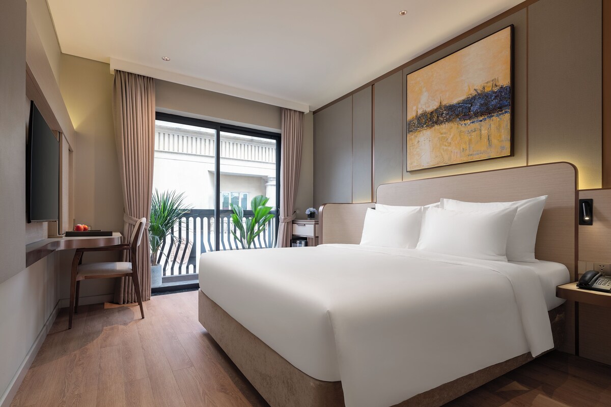 A modern hotel room with a large bed, a desk, a chair, and a view of the outdoors.