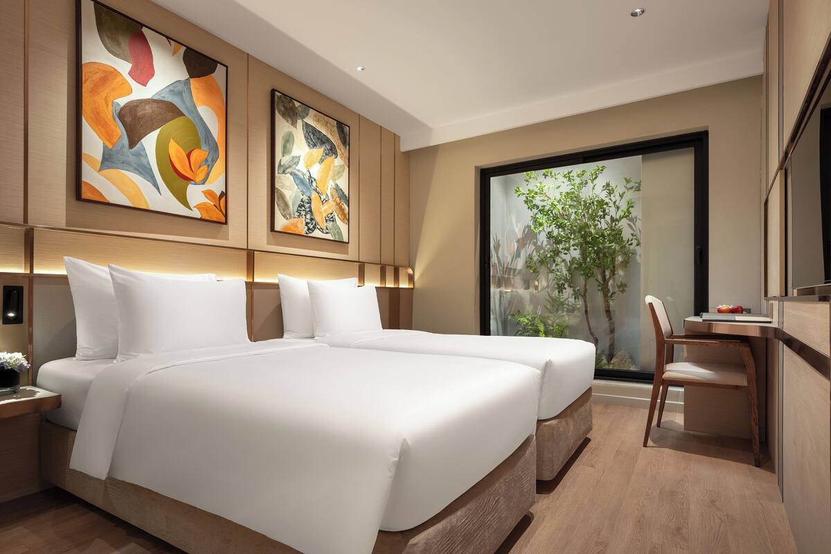 A modern hotel room with a large bed, two bedside tables, a desk with a chair, and large windows with a view of greenery.