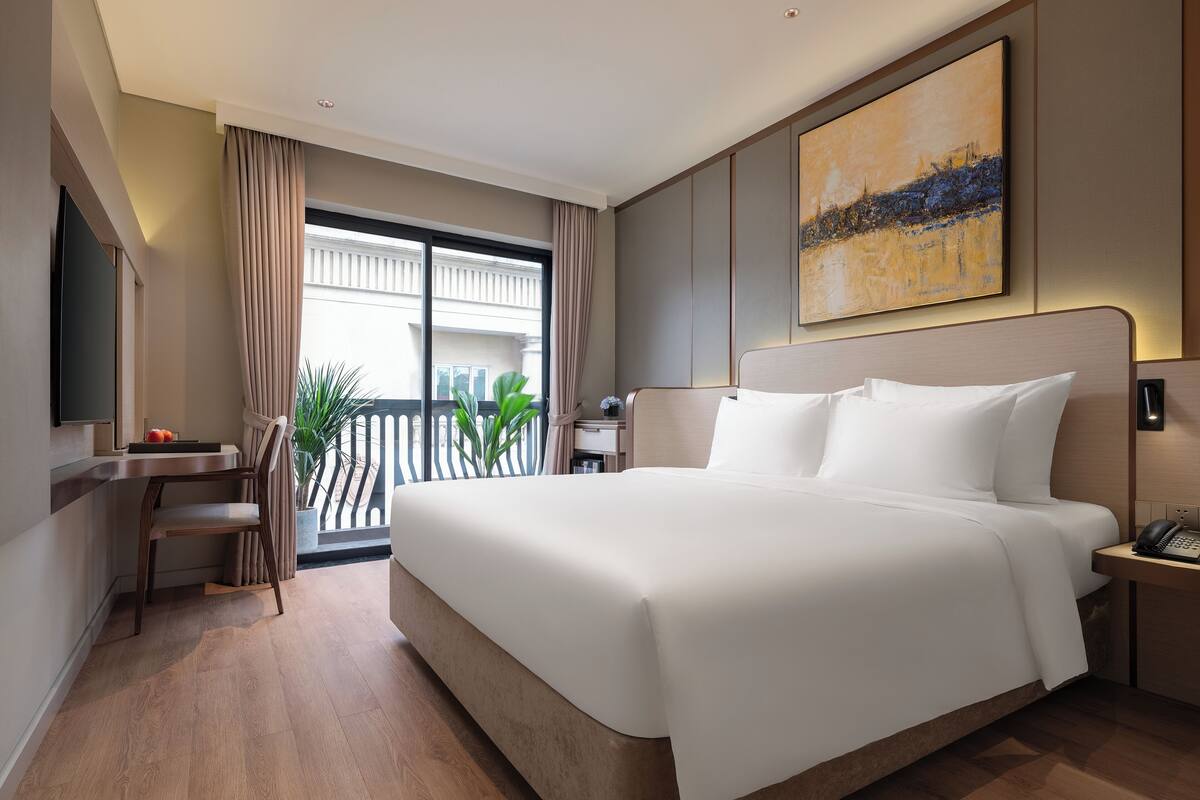 A modern hotel room with a large bed, a desk, a chair, and a view of the outdoors.