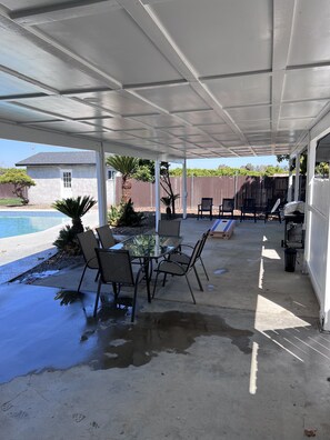 A covered patio with a glass table and chairs, a barbecue grill, and a swimming pool.