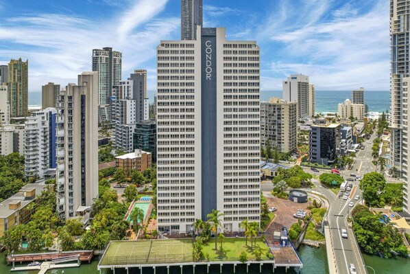 A high-rise building with a prominent 'CONDOR' sign, surrounded by other tall buildings, a marina, and a road with vehicles.