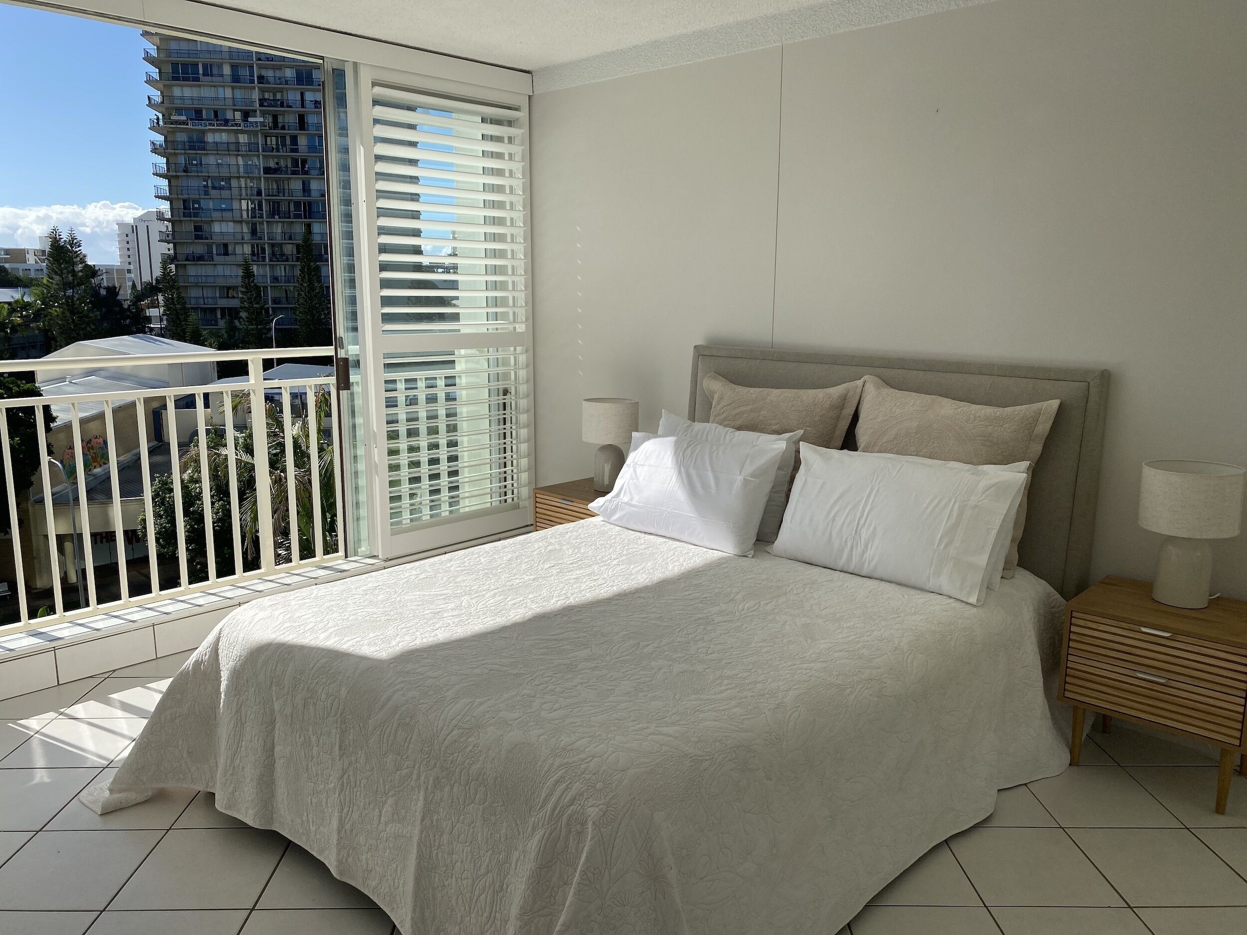 Ein Schlafzimmer mit einem großen Bett, zwei Nachttischen, einem Fenster mit Jalousien und Blick auf ein Gebäude.