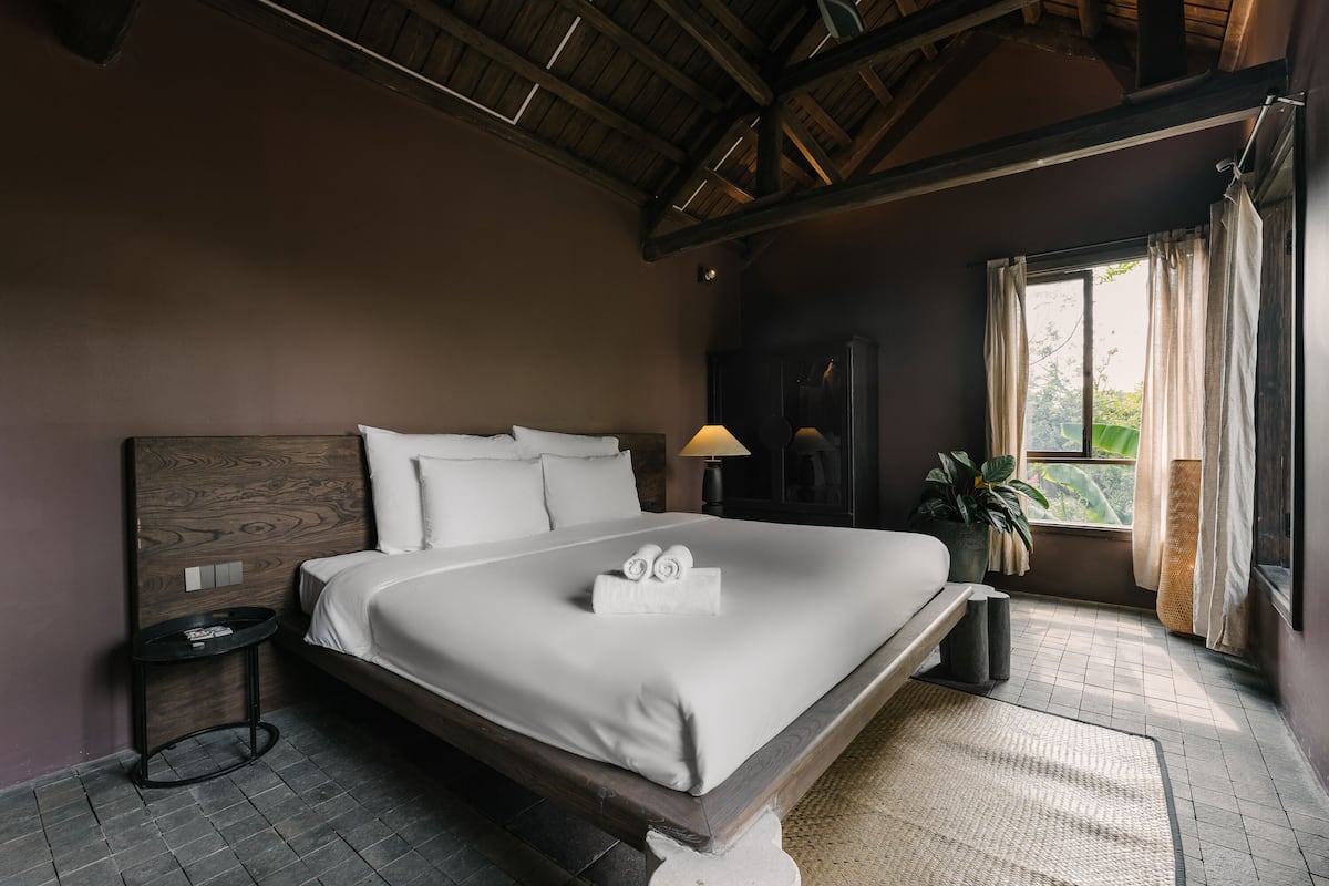 A large bed with a wooden headboard, a bedside table, a lamp, and a window with curtains.