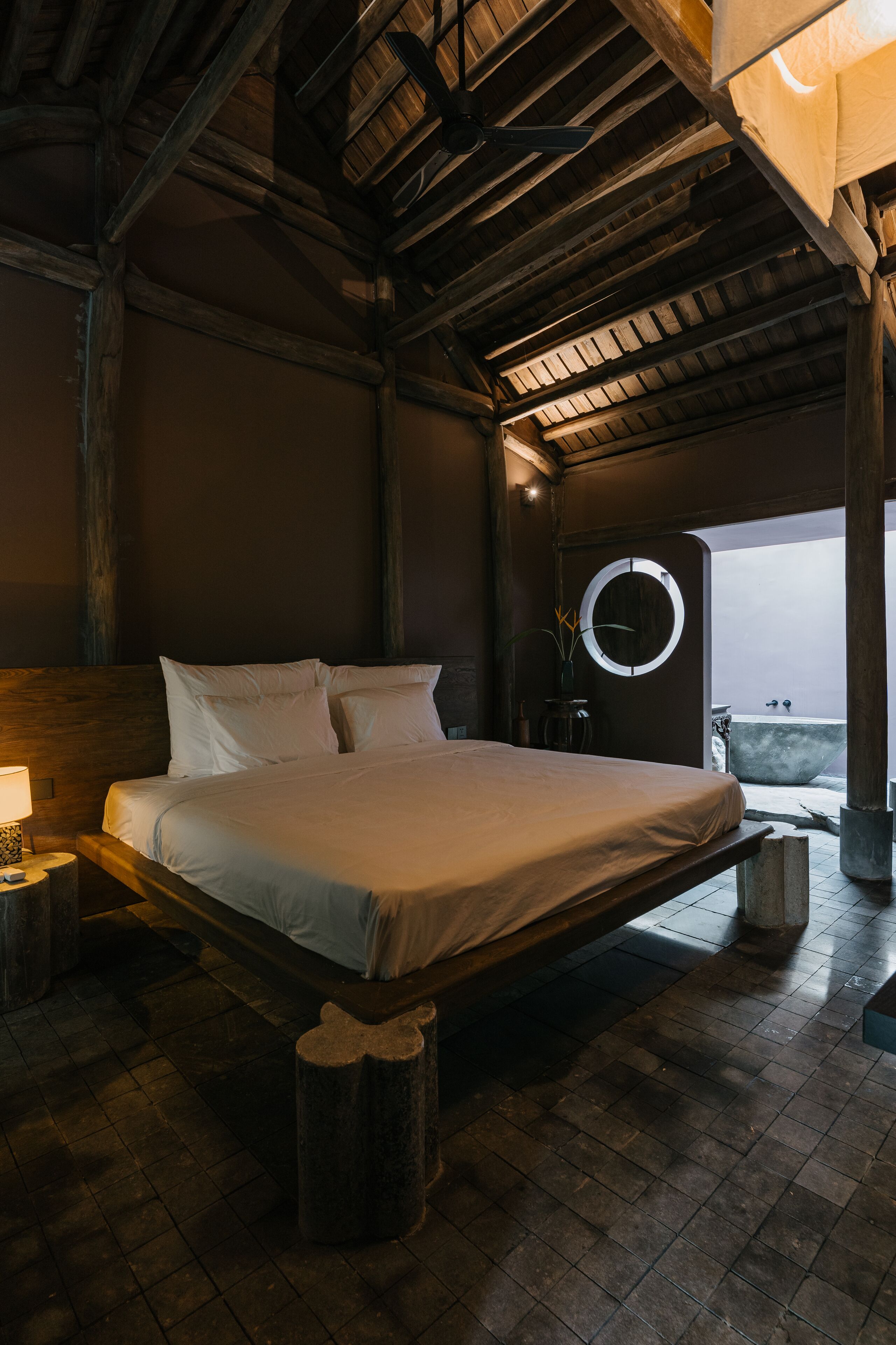 A bedroom with a wooden ceiling, a bed with white linens, a round mirror, and a view of the sea.