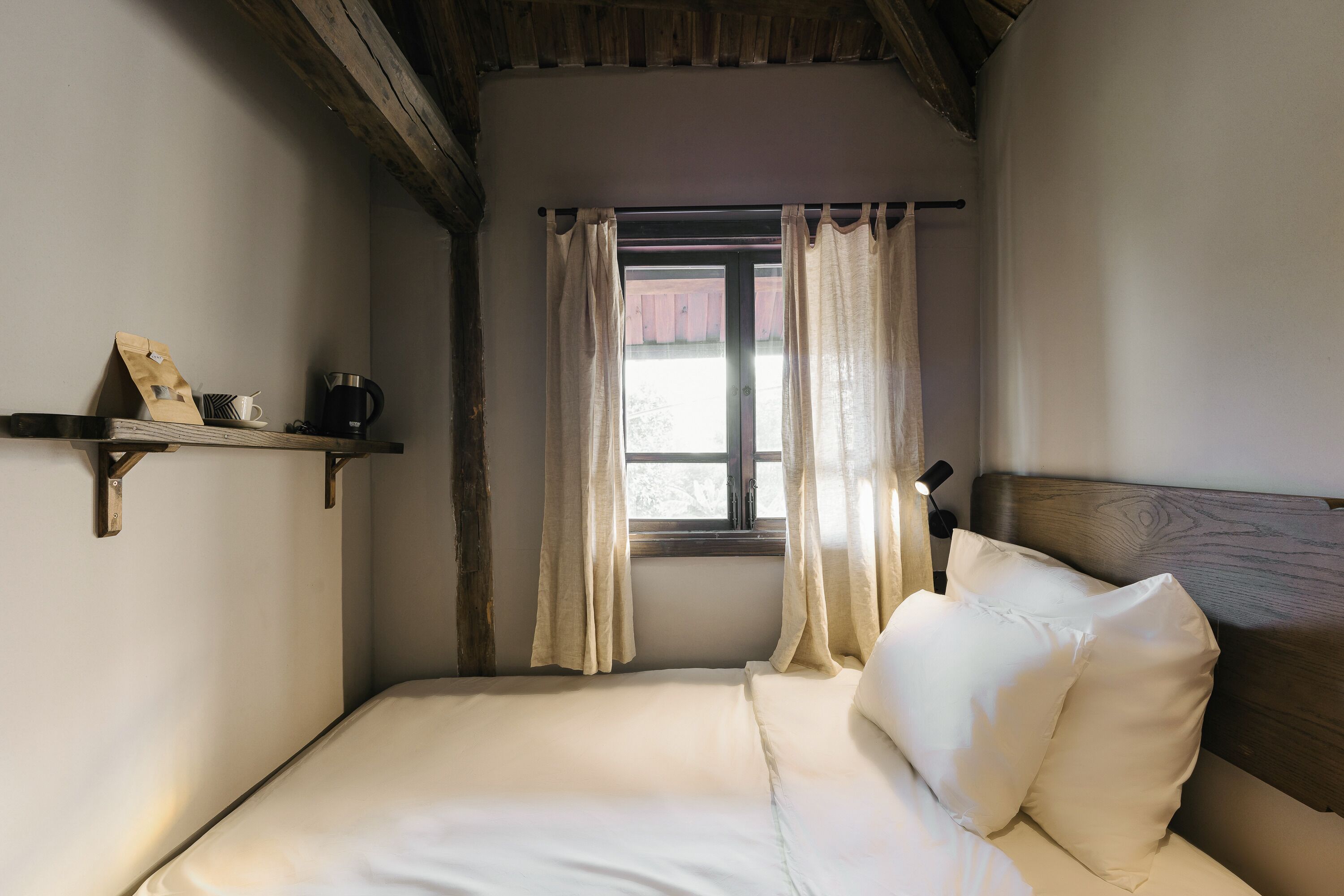 Un dormitorio con cama, almohadas, estante con una taza y un bolso, y una ventana con cortinas.