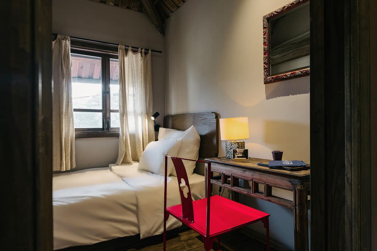 A bedroom with a single bed, a desk with a lamp, a chair, and a window with curtains.