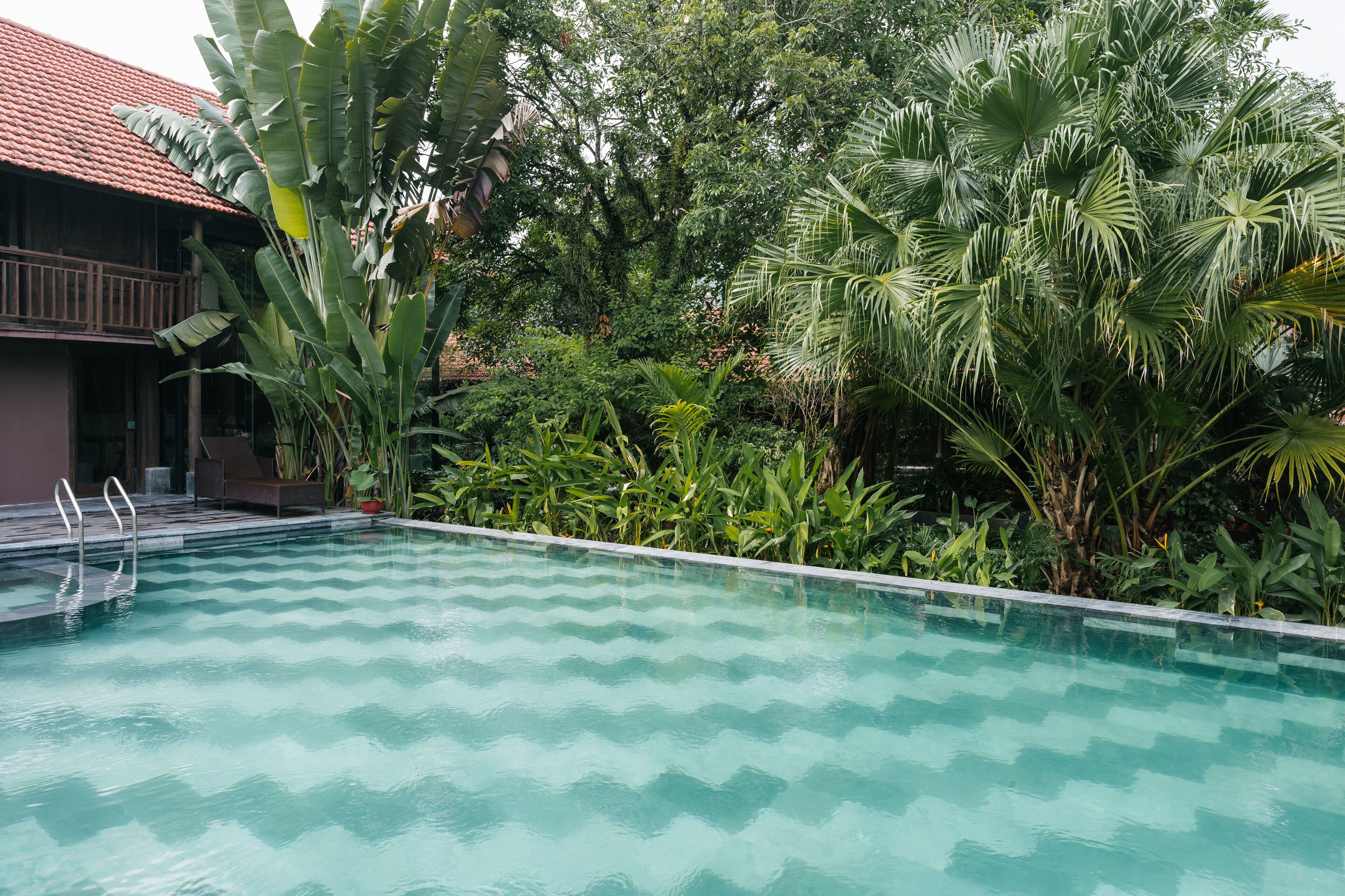 Una piscina con diseño en forma de estampada, rodeada de exuberante vegetación tropical y un edificio con tejado de tejas rojas.