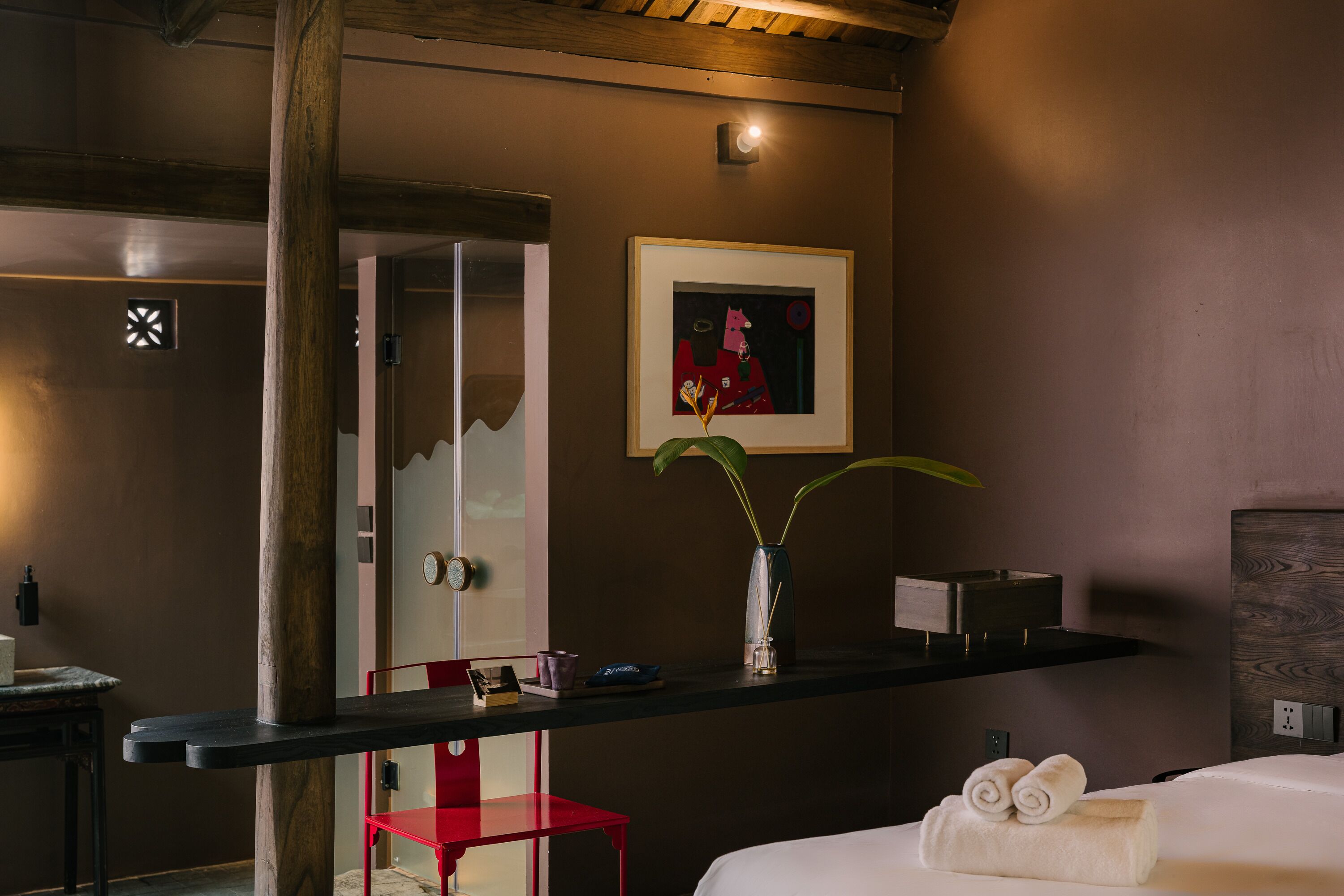 A hotel room with a bed, a red chair, a desk with a vase, and a framed picture on the wall.