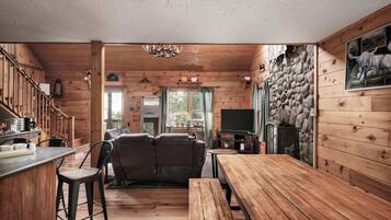 A cozy cabin interior with a wooden dining table, a sofa, and a kitchen area with a stone wall.