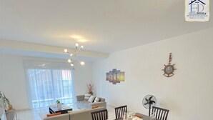 A modern dining area with a large table set for six, surrounded by wooden chairs. A dining table fan and a wall clock are visible. The room has a clean, minimalist design with white walls and a tiled floor. -  E&D Apartment with balcony, Quiet and Secure Ideal For Families (Tananarive)