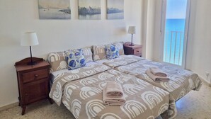 Une chambre à coucher comprenant un lit, deux tables de chevet, une fenêtre donnant sur la mer et trois tableaux accrochés au mur.