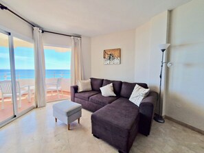 A living room with a dark brown sofa, two white cushions, a grey ottoman, and a floor lamp. A painting hangs on the wall, and a balcony with a view of the sea is visible through the window. - SR Malagueta, Waterfront, Stunning Ocean View (Malaga)