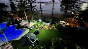 Blick auf einen Poolbereich mit Sonnenschirmen und Liegestühlen, umgeben von Bäumen und mit Blick auf Strand und Stadtlichter in der Ferne.