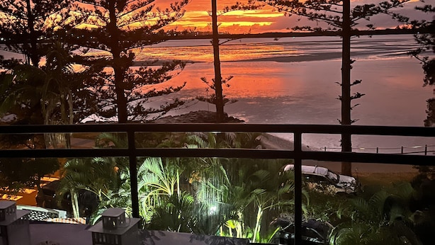 Ein Zimmer mit großem Fenster, das Blick auf einen Sonnenuntergang über einem Gewässer mit Bäumen und Horizont bietet.