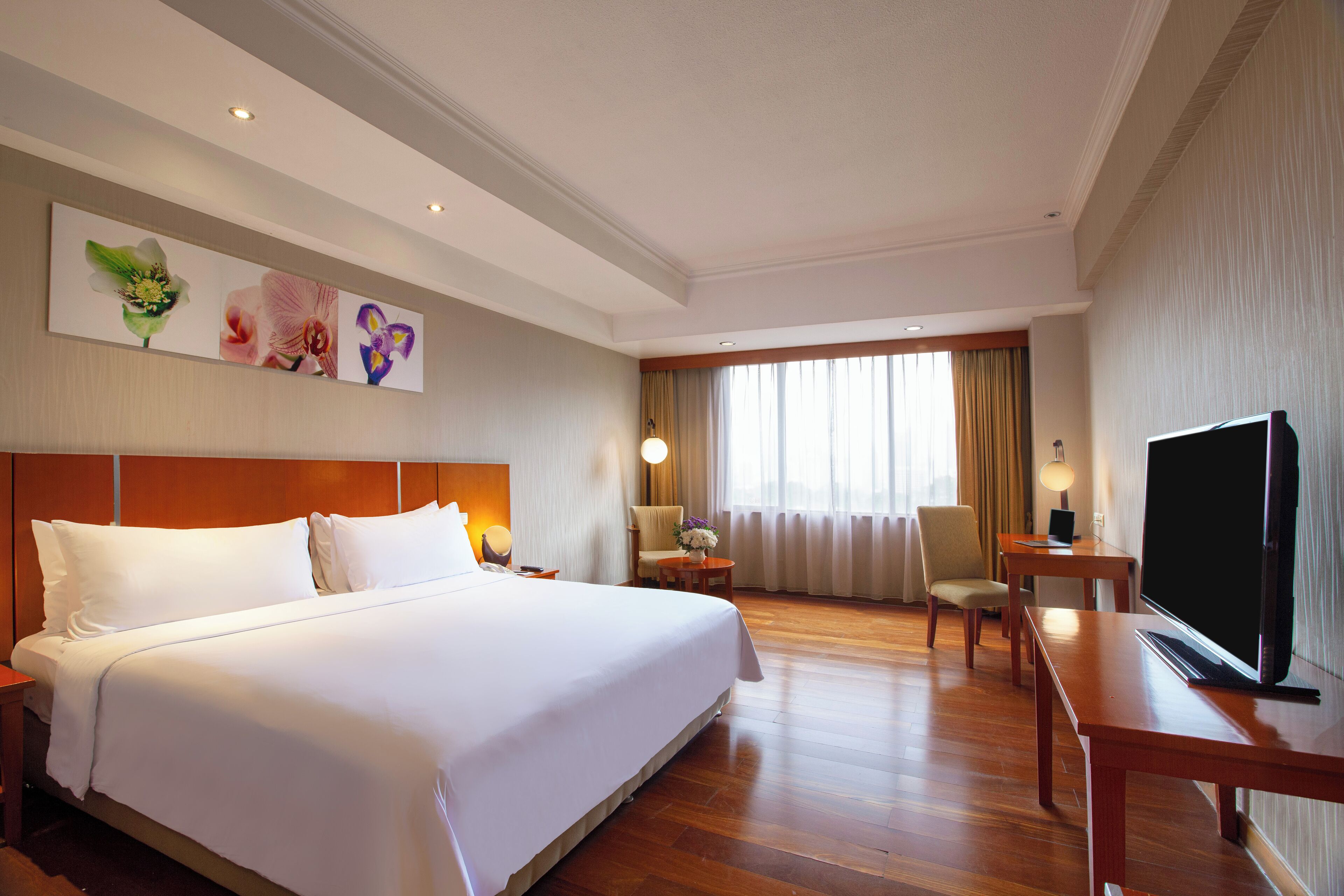 A hotel room with a large bed, a desk with a flat-screen TV, a chair, a small table, and a wall-mounted TV.
