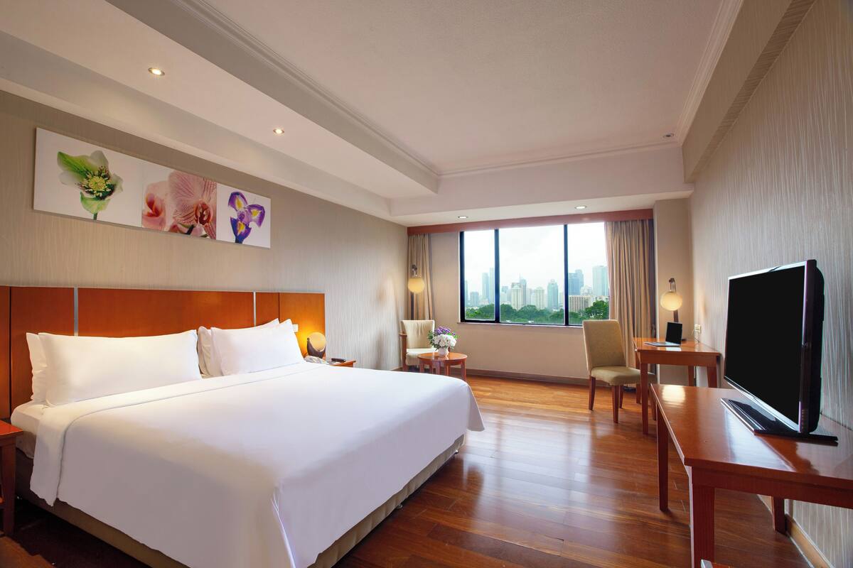 Ein Hotelzimmer mit einem großen Bett, einem Fernseher, einem Schreibtisch und Blick auf die Stadt.