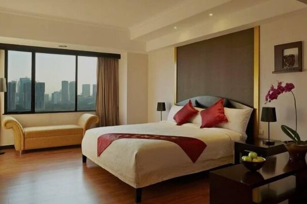 Ein Hotelzimmer mit einem großen Bett, einer Sitzecke, einem Schreibtisch mit Lampe und Blick auf die Skyline der Stadt.