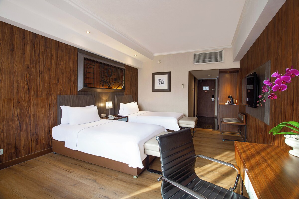 A hotel room with two beds, a wooden floor, a desk with a chair, a TV, and a potted plant.