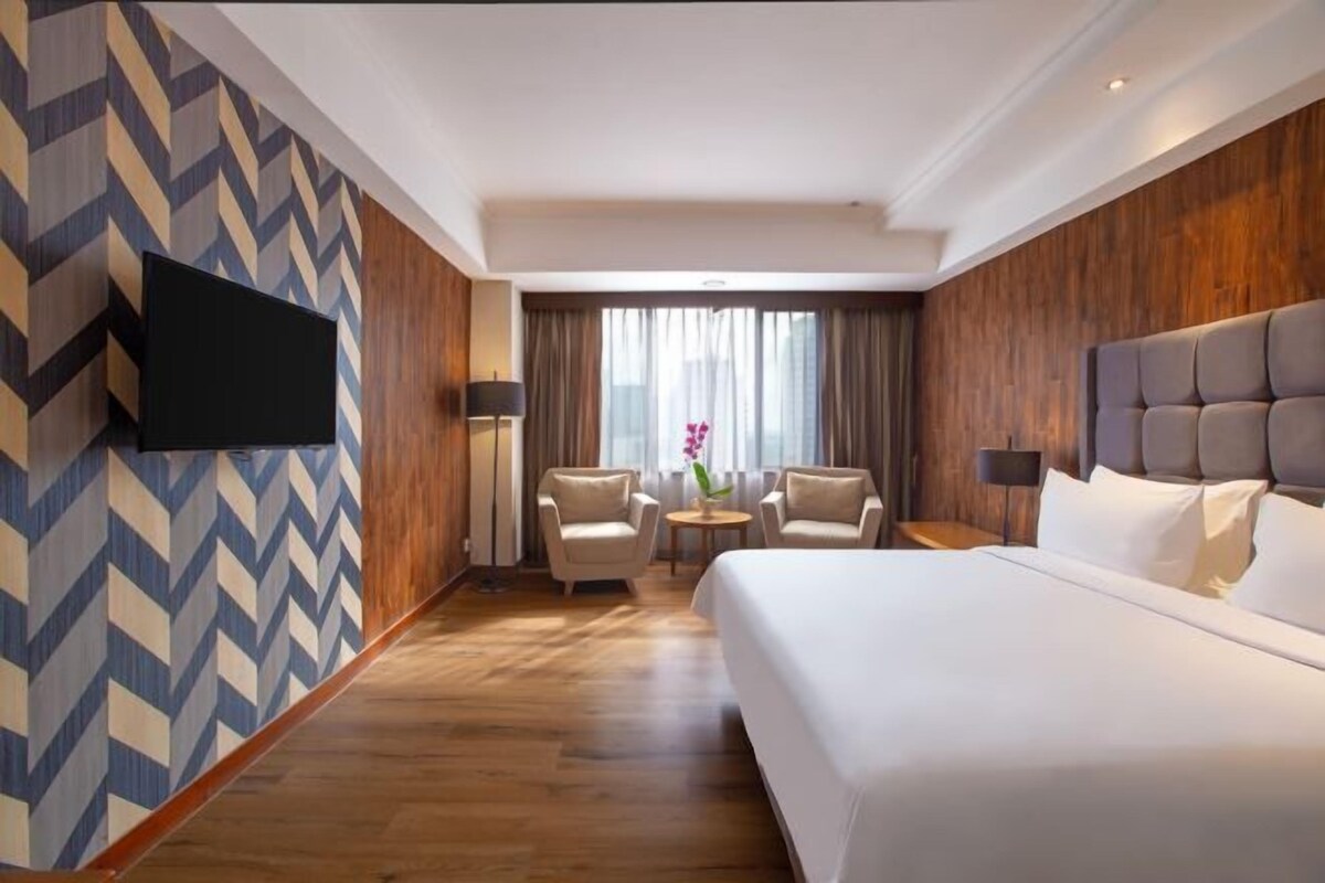 Quarto de hotel com uma cama grande, duas poltronas, uma TV na parede e vista para o exterior através de uma janela.