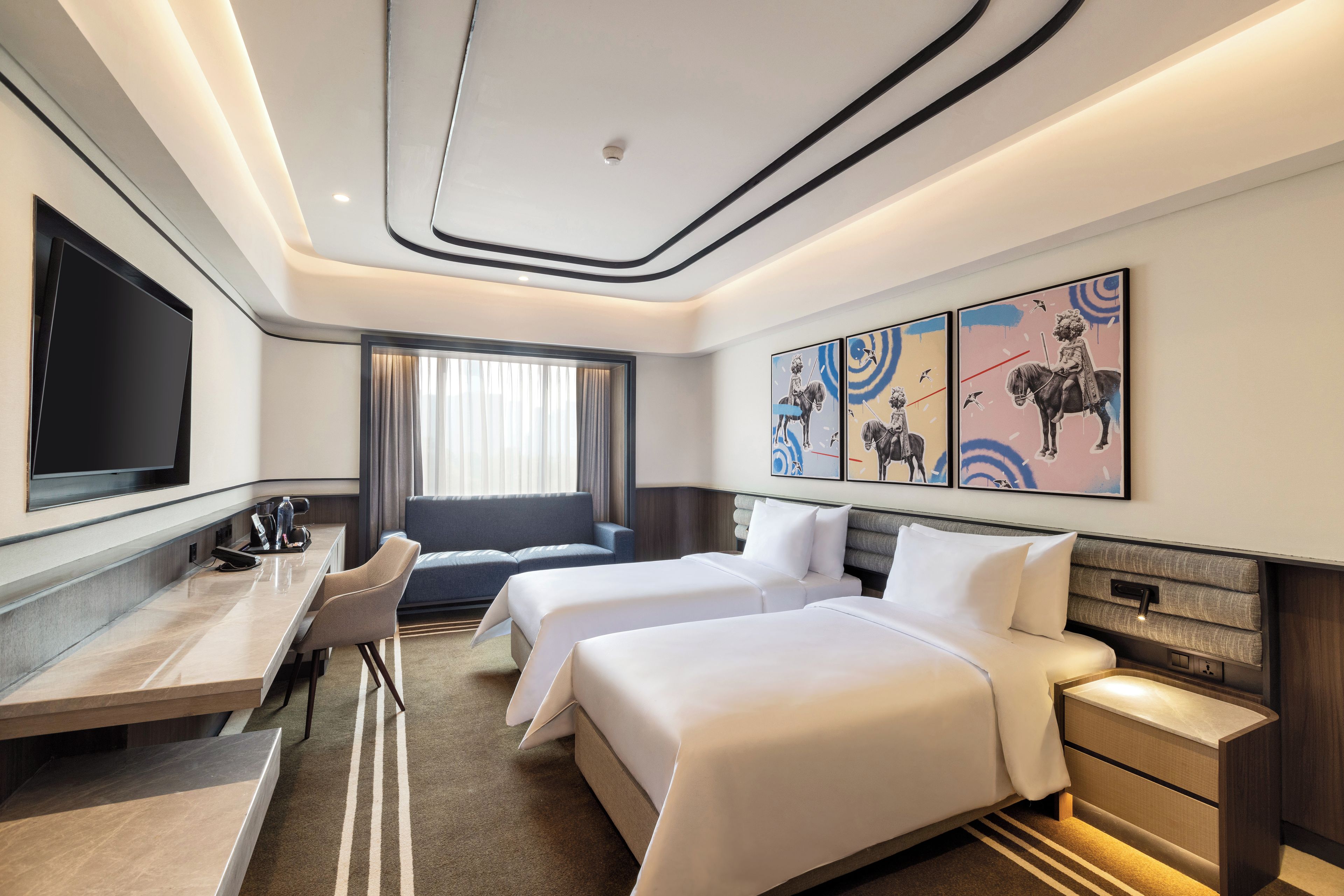 A hotel room with a flat-screen TV, a desk, two beds, a sofa, and artwork on the wall.