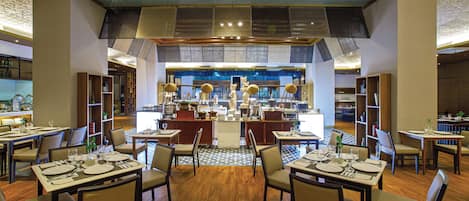 A restaurant with multiple dining tables and chairs, a kitchen in the background, and a patterned ceiling.