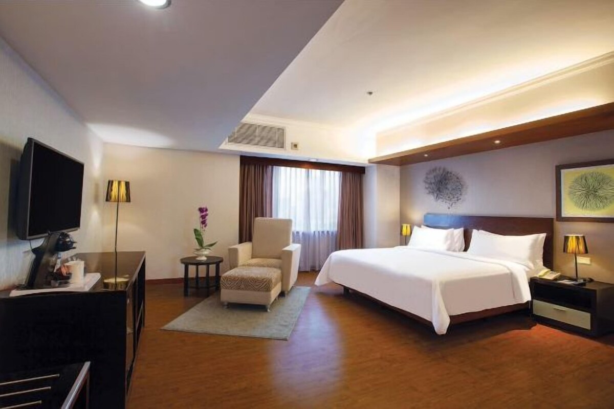 Quarto de hotel com uma cama grande, uma escrivaninha com televisão, uma cadeira e uma pequena mesa com abajur.