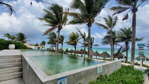 Une piscine à débordement offrant une vue sur l’océan, entourée de palmiers et de parasols en feuilles de palmier.