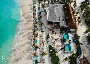 Luchtbeeld van een strandclub met een zwembad, ligbedden en een gebouw met rieten dak.